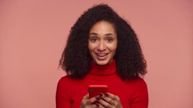 Kırmızı kazaklı, kıvırcık saçlı, gülümseyen ve telefonuna bakarken yumruğunu sıkan pembe arka plandaki başarısını kutlayan bir kadın..