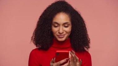 Kıvırcık saçlı, kırmızı kazaklı, neşeli gülümseyen bir kadın. Akıllı telefonunda pembe arka plana karşı olumlu bir mesaj okuyor..