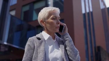 Kendine güvenen, kısa beyaz saçlı, resmi iş kıyafeti giymiş, modern bir ofis binasının dışında telefonla konuşan son sınıf öğrencisi bir kadın..