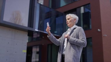 Kısa beyaz saçlı, gülümseyen yaşlı iş kadını akıllı telefondan yapılan video görüşmesi sırasında el sallıyor, modern bir ofis binasının önünde duruyor..