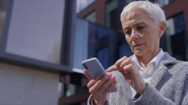 Yetişkin bir iş kadını, kısa beyaz saçlı dokunmatik ekran kullanıyor ve modern bir ofis binasının önünde dururken akıllı telefon içeriğinde kayıyor..