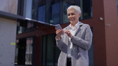 Kısa beyaz saçlı neşeli, olgun bir iş kadını akıllı telefona bakarken iyi haberi ya da başarıyı kutluyor, modern bir ofis binasının önünde duruyor..