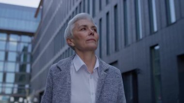 Kısa beyaz saçlı olgun bir kadın modern bir şehir merkezinde yürüyor, hafifçe gülümsüyor ve kendine güveni ve amacı ile ileriye bakıyor..