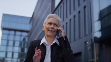 Kısa beyaz saçlı, olgun bir kadın şehir merkezinde yürürken telefonla konuşuyor. Modern iş bölgesinde endişe ya da şaşkınlığını dile getiriyor..