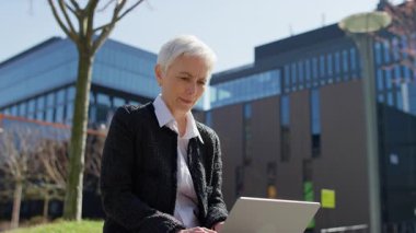 Olgun bir iş kadını, modern bir kentsel alanda dışarıda otururken dizüstü bilgisayar kullanıyor, gün ışığında uzaktan kumandalı işlere odaklanıyor..