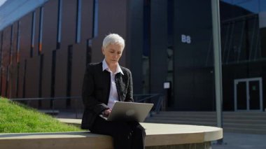 Dışarıda oturan yaşlı iş kadını, bilgisayarında beklenmedik kötü haberleri okurken inançsızlığa ve endişeye kapılıp tepki veriyor..