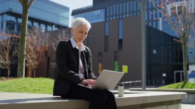 Modern bir şehir ortamında oturup uzaktan çalışma deneyiminin tadını çıkarırken dizüstü bilgisayarda yaşlı bir iş kadını gibi gülümsüyor..