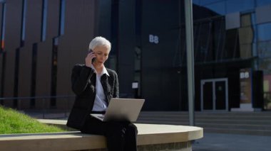 Yaşlı iş kadını dışarıda oturmuş telefonla konuşurken dizüstü bilgisayar kullanıyor, modern şehir ortamında görevleri yönetiyor..