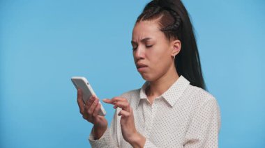 Elinde sıkıntılı bir ifadeyle akıllı telefon tutan genç bir kadın, beklenmedik ya da üzücü haberlere tepki veriyor, bakınırken, mavi bir arka plana karşı.