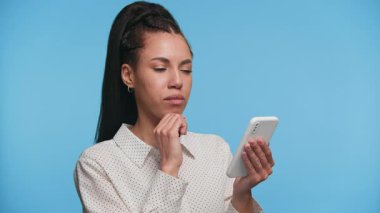 Çevrimiçi alışveriş kararına karşı açık mavi bir arkaplan düşünceli bir ifadeyle akıllı telefon tutan genç bir kadın..