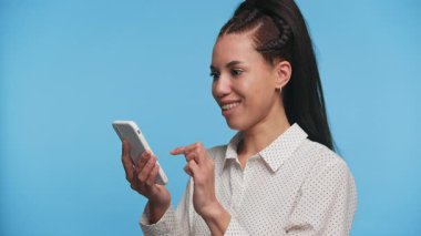 Akıllı bir telefon tutan neşeli genç bir kadın, mavi bir arka plan karşısında, cihazıyla ilgili olumlu ya da heyecan verici haberler karşısında sevinçle tepki veriyor..