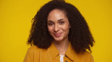 Saf sarı bir arkaplan önünde kıvırcık saçlı, gülümseyen neşeli bir kadının yakın plan portresi..