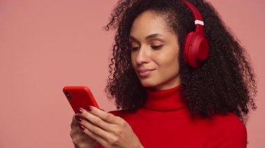Kırmızı kazaklı ve kulaklıklı gülümseyen genç bir kadın akıllı telefonuna pembe bir arka plana mesaj yazıyor..