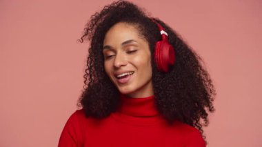 Pembe arka planda kulaklıkla müzik dinlerken gözleri kapalı, rahat genç bir kadın gülümsüyor..