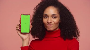 Kadın yeşil ekran maketli akıllı telefonu gösterirken bunu onaylamıyor, pembe arka plandaki memnuniyetsizliği veya anlaşmazlığı dile getiriyor.