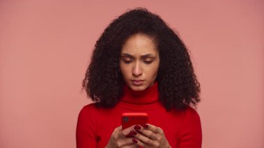 Kırmızı kazaklı kadın elinde akıllı telefon tutarken ve pembe arka planda şok edici bir şey okurken inançsızlıkla tepki verirken....