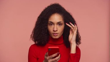Kaygılı, kıvırcık saçlı, elinde telefon olan genç bir kadın. Ekranda pembe arka plana karşı üzücü bir şeyler okuyor..