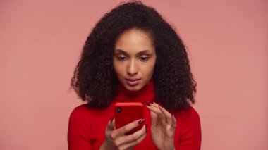 Kırmızı kazaklı stresli genç bir kadın endişeli bir ifadeyle akıllı telefon tutuyor. Pembe arka planda belaya veya kötü habere tepki veriyor..
