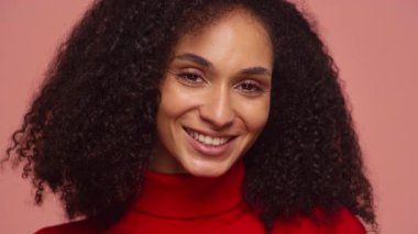 Kıvırcık saçlı neşeli genç bir kadının portresi. Kırmızı balıkçı yakalı, yumuşak pembe bir arka planda güvenle gülümsüyor..