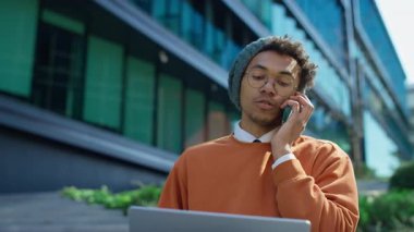 Modern öğrenci ya da serbest çalışan bilgisayar ve akıllı telefonla dışarıda çalışan, cam binanın yanındaki merdivenlerde oturan, bir telefon görüşmesinde konuşan.