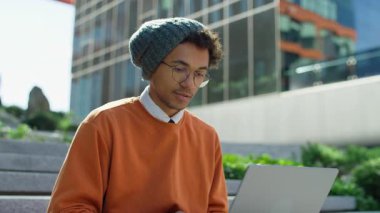 Genç bir erkek öğrenci, modern bir üniversite binasının merdivenlerinde otururken dizüstü bilgisayarında gözlük ve bere takıyor..