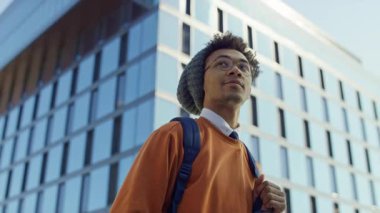 Sırt çantalı genç adam, öğrenci hayatı ve akademik yolculuğu temsil eden modern cam bir binanın dışında güvenle yürüyor..