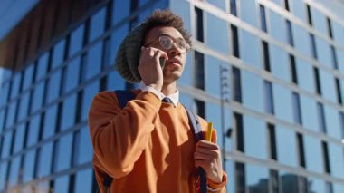 Sırt çantası ve not defteri olan genç bir öğrenci modern cam bir üniversite binasının önünde güpegündüz yürürken akıllı telefondan konuşuyor..
