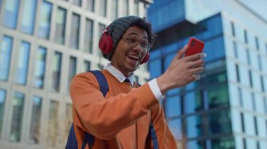 Kulaklık takan ve sırt çantalı genç bir öğrenci, modern kampüs binalarının dışında yürürken akıllı telefon kullanarak bir video görüşmesinde konuşuyor..
