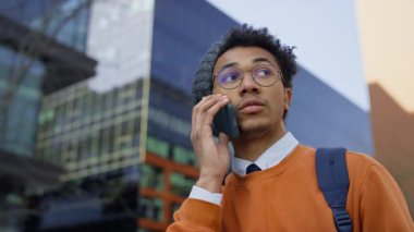 Sırt çantası ve gözlüklü genç erkek öğrenci modern kentsel binaların yanında yürürken ciddi bir telefon konuşması yapıyor..
