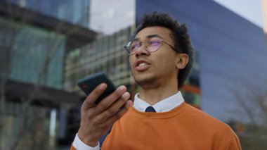 Turuncu kazaklı şık genç adam modern cam binaların dışında sesli mesaj kaydederken elinde akıllı telefon tutuyor..