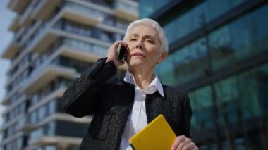 Yetişkin, beyaz saçlı, kısa boylu, profesyonel bir kadın, elinde ofis binasının yanında sarı bir not defteri tutarken dışarıda telefon görüşmesi yapıyor..