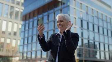 Akıllı telefondan yapılan video görüşmesi sırasında gülümseyen yaşlı iş kadını, modern ofis binasının yakınında gündüz vakti duran....