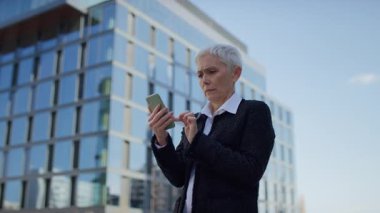 Üst düzey kadın, elinde akıllı telefon tutarken, sıkıntılı yüzünü kapatıyor. Bu da modern binaların yakınında kötü haber veya duygusal tepkiyi işaret ediyor..