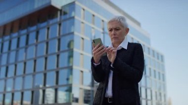 Yaşlı kadın endişeli bir ifadeyle akıllı telefona bakıyor, rahatsız edici haberlere tepki veriyor veya şehir dışındaki beklenmedik ihbarlara tepki veriyor.