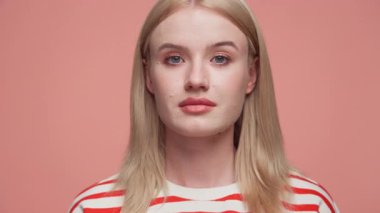 Düz sarı saçlı, çizgili gömlekli, yumuşak pembe arka planlı, ciddi bir genç kadının yakın plan portresi..