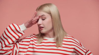 Kırmızı çizgili tişörtlü sarışın kadın hayal kırıklığını ya da stresi gösteriyor. Alnına pembe arka planda kapalı gözlerle dokunuyor..