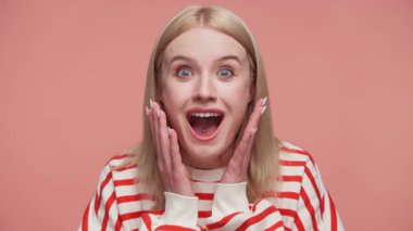 Elleri yanaklarında, geniş gülüşlü, neşeli bir şok ve pembe bir arka planda heyecan ifade eden sarışın kadın..