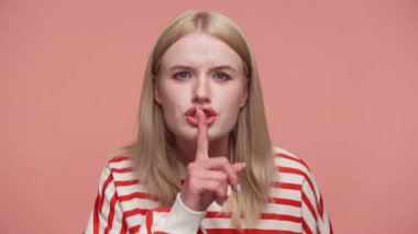 Çizgili tişörtlü sarışın kadın parmaklarını dudaklarına uzatarak sessiz bir jest yapıyor. Pembe arka planda odaklanmış bir ifadeyle sessizliği işaret ediyor..