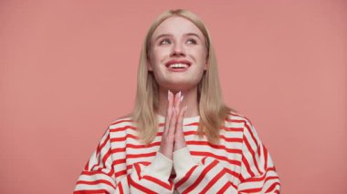 Çizgili gömlekli sarışın kadın ellerini sıkarak yukarı bakıyor ve gülümsüyor, umut dolu arzusunu dile getiriyor ya da pembe arka planda bir dilek diliyor..