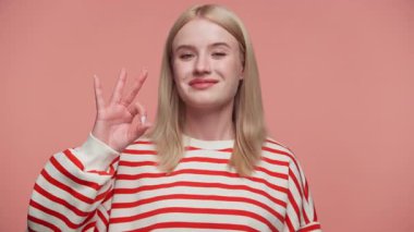 Kırmızı çizgili tişörtlü sarışın kadın kendine güvenerek gülümsüyor ve stüdyoda pembe yumuşak bir arka planda el işareti gösteriyor..