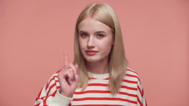 Kırmızı çizgili tişörtlü sarışın kadın ciddi bir yüzle işaret parmağını kaldırıyor. Pembe arka plana karşı uyarı ya da onaylamama işareti yapıyor..