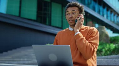 Kendine güvenen genç girişimci dışarıda dizüstü bilgisayarla oturmuş modern cam ofis binasının yakınındaki bir telefon görüşmesinde konuşuyor..