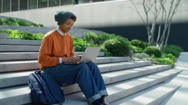 Şehir merdivenlerinde dizüstü bilgisayarla oturan, yeşillik ve modern binaların yakınında çalışan gündelik kıyafetli genç girişimci..