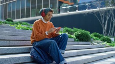 Endişeli öğrenci dışarıda merdivenlerde oturuyor, kulaklık takıyor ve modern kampüs binasının yakınında endişeli bir ifadeyle akıllı telefona bakıyor..
