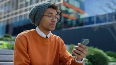 Modern bir binanın önünde oturan öğrenci, akıllı telefonuyla yaptığı bir video konferans sırasında, hayal kırıklığıyla el kol hareketi yapıyor..