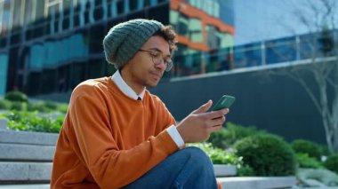 Genç adam modern binanın yakınındaki merdivenlerde oturuyor. Sıkılmış ve ilgisiz bir ifadeyle akıllı telefonunu karıştırıyor..