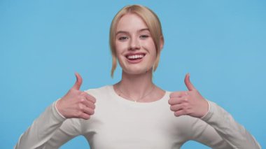 Beyaz bluzlu neşeli genç bir kadın, parlak mavi bir arka plana karşı duruyor, kendine güvenerek gülümsüyor ve onaylamak için çifte başparmak işareti yapıyor..