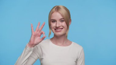 Beyaz tişörtlü, gülümseyen genç bir kadın parlak mavi bir stüdyo arka planına karşı eliyle iyi bir işaret veriyor..
