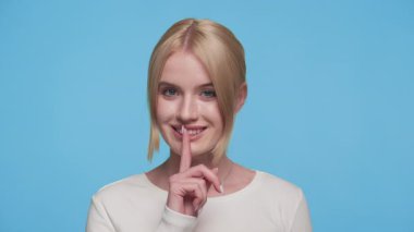 Gülümseyen genç bir kadın, dudaklarında parmaklarıyla sessizlik işareti veriyor, beyaz bluz giyiyor, parlak mavi bir arka plana karşı, neşeli bir ifade..