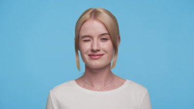 Gülümseyen genç bir kadının portresi, tek bir göz kırpma yapıyor, mavi bir arkaplanda duruyor, sıradan ve eğlenceli bir ifadeyle..